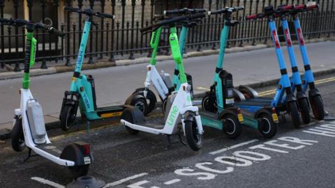 A row of electric scooters from various brands, including Lime, TIER, and Dott, are parked neatly in a designated "E-scooter hire" zone on the side of a city street. The scooters are lined up beside a black iron fence and a pavement.