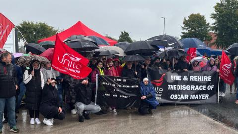 Striking workers stand together in the rain holding banners saying "Fire and rehire the disgrace of Oscar Mayor" and Unite flags