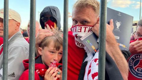 A man stands with his arms around a young boy who looks distressed. The man holds a Liverpool Football scarf around his nose and mouth and they are surrounded by other fans. They are standing behind metal bars. 