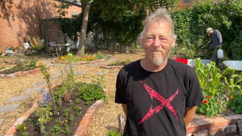 A man with half a beard, grey hair and a black t-shirt with a red 'X' printed on it, stands in a garden. The garden has beds of plants made with bricks and old filing cabinets. There are also paving slabs amongst the wood chipping garden surface, a scarecrow and a large tree. A man in a grey jumper and olive bucket hat is watering the plants. There are brick walls bordering the garden, partially covered by leaves.