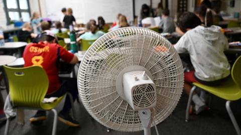 A picture taken from the back of a classroom showing a large white fan blowing air towards young children studying at their desks