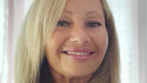 Head shot photograph of a woman with a blonde shoulder length hair, smiling at the camera