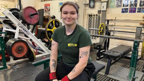 Charlotte Macaulay, who wears a green weightlifting club t-shirt and has tattooed arms, smiles while sat on a bench in the gym 