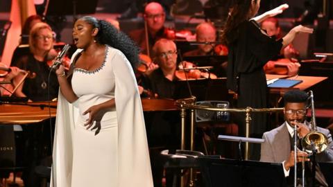 Samara Joy, wearing white and singing into a microphone, on stage at the Proms with musicians around her.