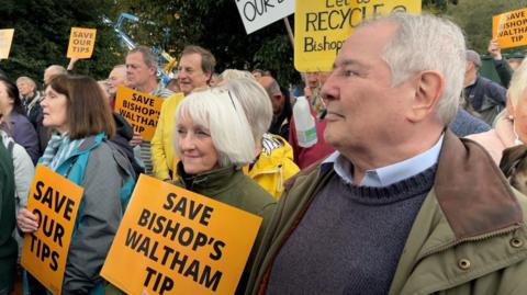 A crowd of people face to the left of the camera. Some hold orange signs with the words "Save Bishop's Waltham tip"