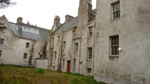 A derelict white building with a grassy area to the front