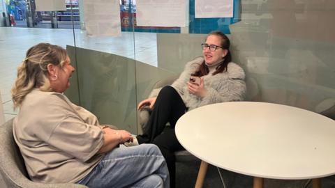 18-year-old Demi sitting inside the Epic Youth Hub chatting to one of the members of staff there. Demi is holidng her phone in her left hand and both her and the member of staff are smiling at each-other.