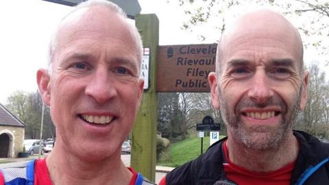 A man with short grey hair stood next to a man with a shaved head and stubble. Behind them a sign says Cleveland Way, Rievaulx and Filey.