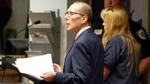 Sheila Keen-Warren and her lawyer stand together in front of a microphone in a courtroom. He wears a suit and glasses as he holds some papers. She has long blonde hair and wears a navy jumpsuit.