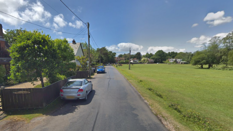 Two cars are parked outside houses on Meerut Road in Brockenhurst, opposite a grassy common.