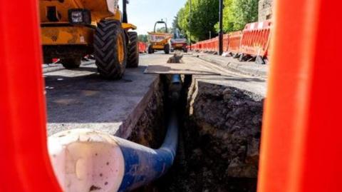 Work being carried out on a road in Bradford, there is a water pipe visible and several diggers. There are red plastic barriers on each side.