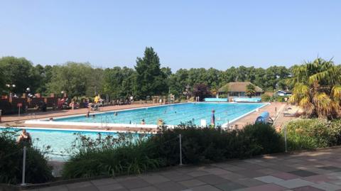 The outdoor pool seen from the far end. There is a small shallow pool and a 50-metre big pool with swimming lanes. There is a lifeguard seat on the left, with one or two lifeguards pictured wearing yellow and red uniform. There are palm trees and other trees around the sides.