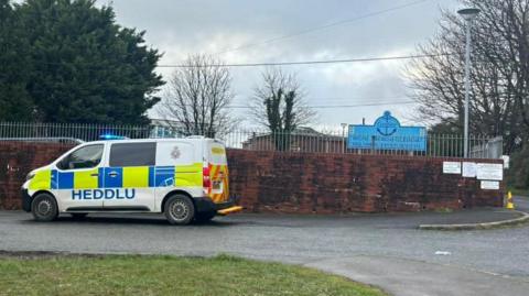 A brown school wall with a silver railing above and a police van parked outside. In the background is a blue school sign.
