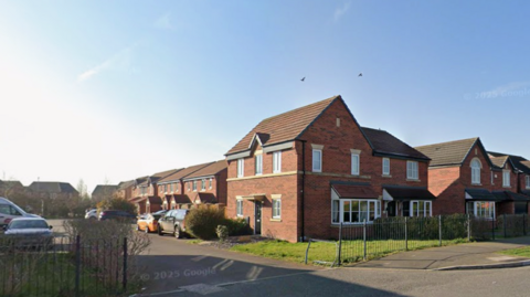 The opening of a road with houses and cars along it. They are set against a blue sky, and there is railing along a pavement in the foreground of the image.