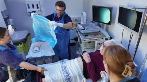 A man laying on a hospital bed with a medical sheet across his lower abdomen and thighs. Three members of medical staff are standing around him - two women and a man. There are monitor screens above his head and medical kit around him