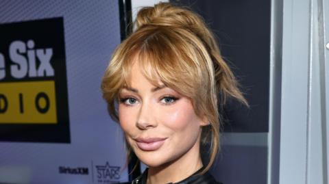 A woman smiles at the camera. She has blonde hair that has been tied back and bangs frame her face. 