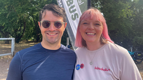 A man and a woman standing together in front of a green and white banner that reads "bike bus". The man has brown hair and is wearing sunglasses and a blue t-shirt. The woman has pink and brown hair and is wearing a light pink t-shirt