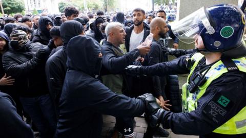 Police officers stand guard as a large group of angry-looking counter-protesters push against the police cordon on the day of a protest against illegal immigration