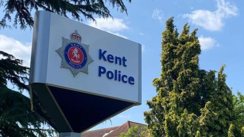 A white sign with the words Kent Police written on it in blue, with the Kent Police logo to the left. Behind the sign are green trees