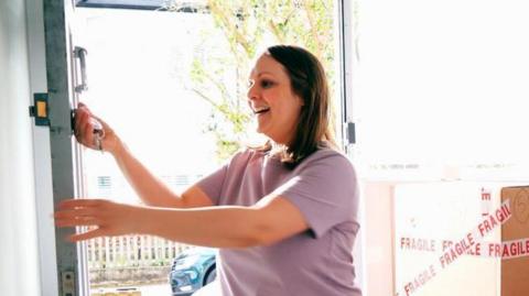 A woman looks happy with her key in the lock of an open front door of a house, as though she is moving in. She has shoulder-length dark hair and wear's a light purple T-shirt. A cardboard box with the words "fragile" in tape across it is next to get