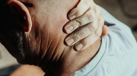 A bald man, pictured from behind with his hand clenched over the back of his neck, as if it is in pain.