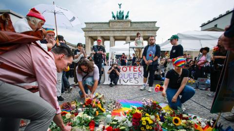 People lay flowers in front of a banner saying 'protect queer lives', with the Brandenburg Gate in the background on a cloudy day in Berlin on Sunday. 