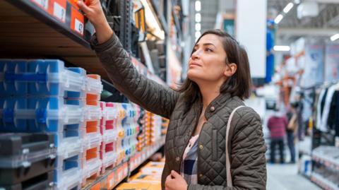 A woman with dark brown hair, wearing a green coat, shopping in a construction store. She is reaching up at a shelve which contains boxes.