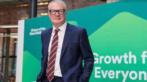 Man with white hair standing in a navy blue suit, in front of a green sign saying growth