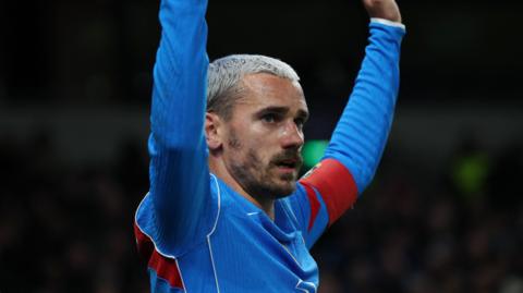 Atletico Madrid forward Antoine Griezmann, with bleached blond hair and facial hair, has both arms raised as he wears blue and red kit