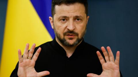 Volodymyr Zelensky in front of a Ukrainian flag with his hands up