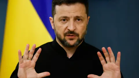 Volodymyr Zelensky in front of a Ukrainian flag with his hands up
