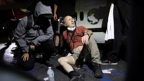 A man wearing a t-shirt, trousers and trainers sits on the pavement, one trouser leg rolled up showing gauze wrapped around his right thigh. He wears a camera around his neck and a respirator mask pulled down. Two people look on to him. 