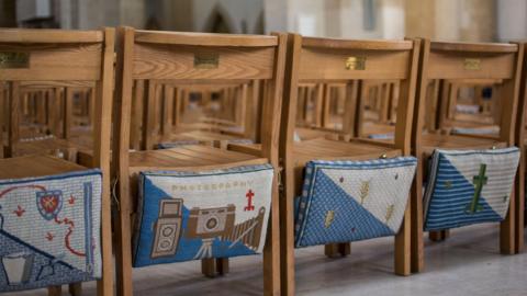 Four church kneeling cushions hanging off the back of chairs. They feature artwork including gardening, a camera, church motifs and a green cross.