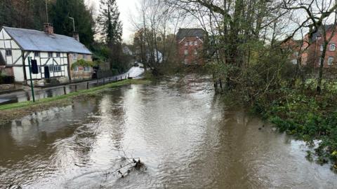 A very swollen river with a road to the left of the image where a large amount of water can still be seen, without houses in the background and a few bare trees