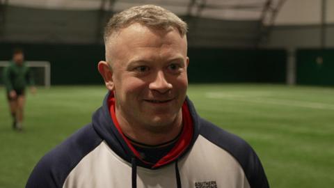 Dan Robinson is a white man, wearing a white hoody, with blue sleeves and good, he has cropped grey hair and is smiling. He's standing on a green all-weather pitch where some of his fellow rugby players can be seen warming-up in the background.