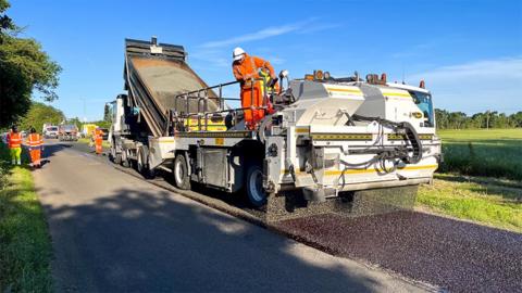 A lorry deposits stone chippings on a road while another machine flattens it into the bitumen