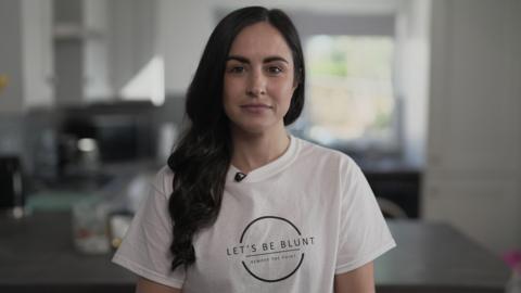Leanne Lucas, who has long black hair, looks into the camera while wearing a white t-shirt with a circular logo with the words 'Let's Be Blunt' printed in black lettering