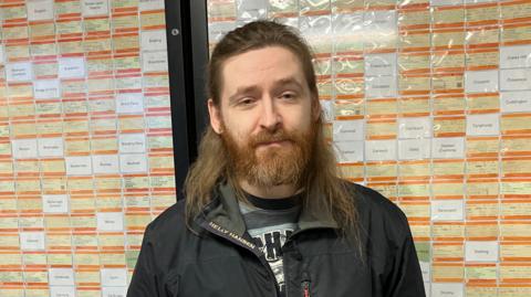 Elliott Badger who has should-length brown hair pulled back from his forehead and a beard. He is wearing a black jacket and standing in front of a notice board which is packed with railway tickets. There are also white cards with the type-written names of missing stations