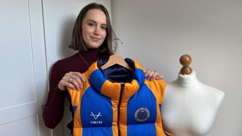 Ms Herman is standing in front of a white wardrobe. She has a brown bob haircut and is holding a blue and amber puffer gilet with the Shrewsbury Town football club logo on it