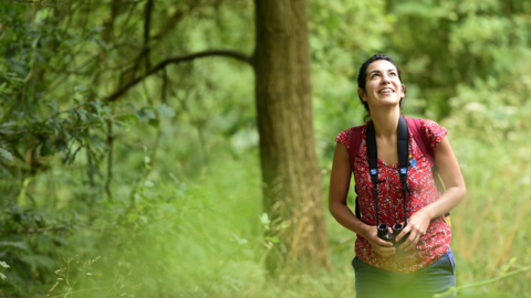 A woman with dark brown hair tied back in a bun is walking through woodland, she is smiling looking towards the sky, wearing a short sleeved red floral print t shirt and blue trousers, a red rucksack and carries binoculars in her hands, in the background is a large tree and she is surrounded by greenery while she enjoys birdwatching