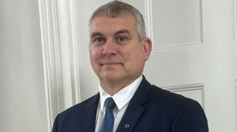 Smith is wearing a dark suit with shirt and tie and is looking at the camera. He has grey and dark hair.