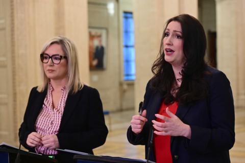 A woman with blonde hair, wearing glasses, a black jacket and a pink shirt, standing beside a woman with brown hair, wearing a black jacket and red shirt