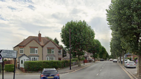 A residential road with houses on either side, parked cars, and a road sign pointing toward a nearby school and community centre.
