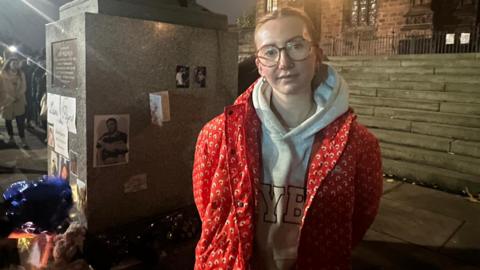 A woman with hair tied back, wearing glasses, a grey hoody and a red coat, stands in front of the statue, where tributes to Liam Payne have been laid.