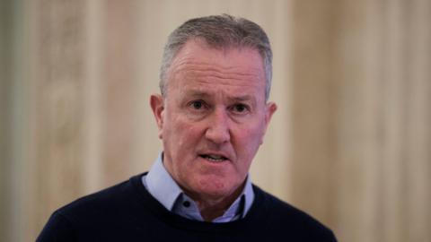 Conor Murphy, grey hair, blue shirt with dark sweater. In Stormont hall