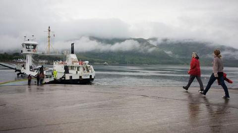 Two women walk towards the ferry, MV Corran. It is a wet day and the jetty is damp, and mist covers hills in the distance.