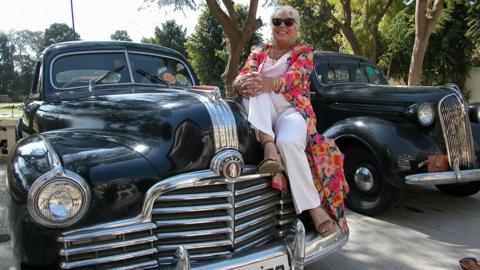 A woman wearing sunglasses and dressed in white with a long, flowing, ankle-length floral overshirt with bright, bold, orange and pink flowers on it sits on the bonnet of a vintage black car with a large chrome grille smiling in the sunshine
