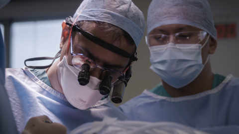 Two people wearing blue scrubs, hants and face masks are carrying out a surgery. They are wearing glasses, one has specialised magnifying equipment - like a tiny telescope, on each lense. 
