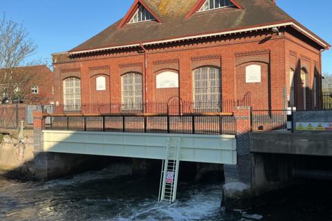A red-brick Victorian pumping station sits behind a new footbridge with metal railings and a cream-coloured structure underneath, with water flowing through
