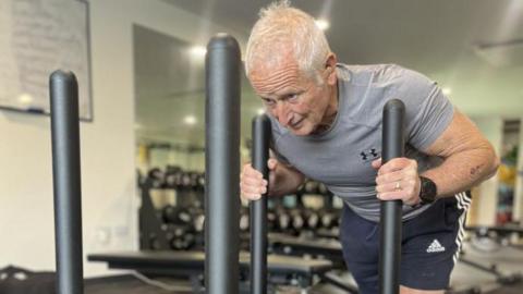 Peter Herbert in a grey tight-fitted shirt and blue shorts. His arms are grasping two vertical bars on an exercise sled which are almost touching his shoulders. His head is down and he is straining in his face and neck. He has white hair. 